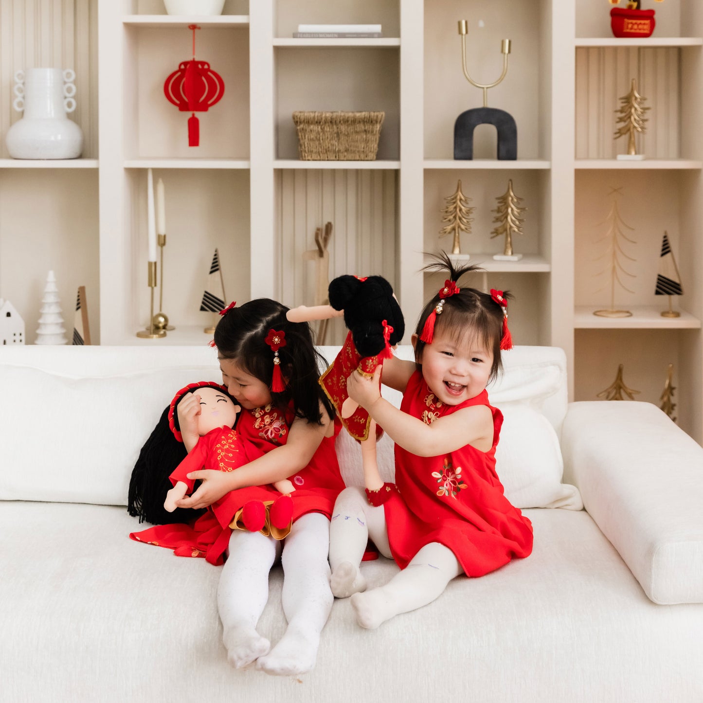 Two young girls laughing joyfully while sitting on a white sofa and playing with their Mei Chinese cultural dolls and Hoa Vietnamese doll. Both girls and their dolls are dressed in matching vibrant red outfits with golden floral embroidery and red floral hair tassels.