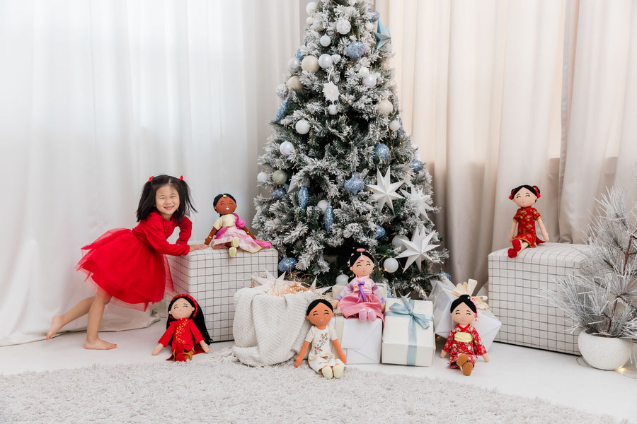 A smiling child in a red dress beside a frosted holiday tree, surrounded by wrapped gifts and Joeydolls cultural dolls