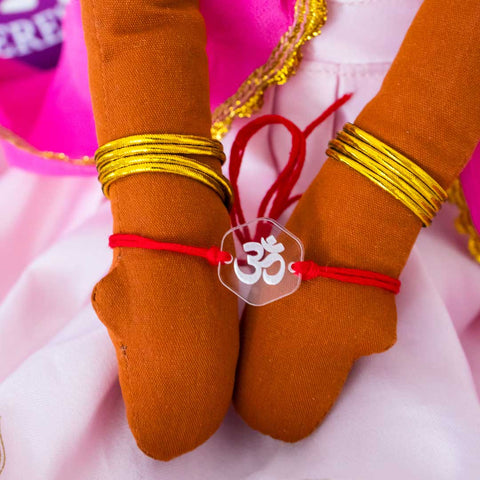 Om Rakhi by Creative Crafty Mom featuring a handmade red thread bracelet with a clear acrylic charm engraved with the Om symbol, tied on a doll’s wrist.