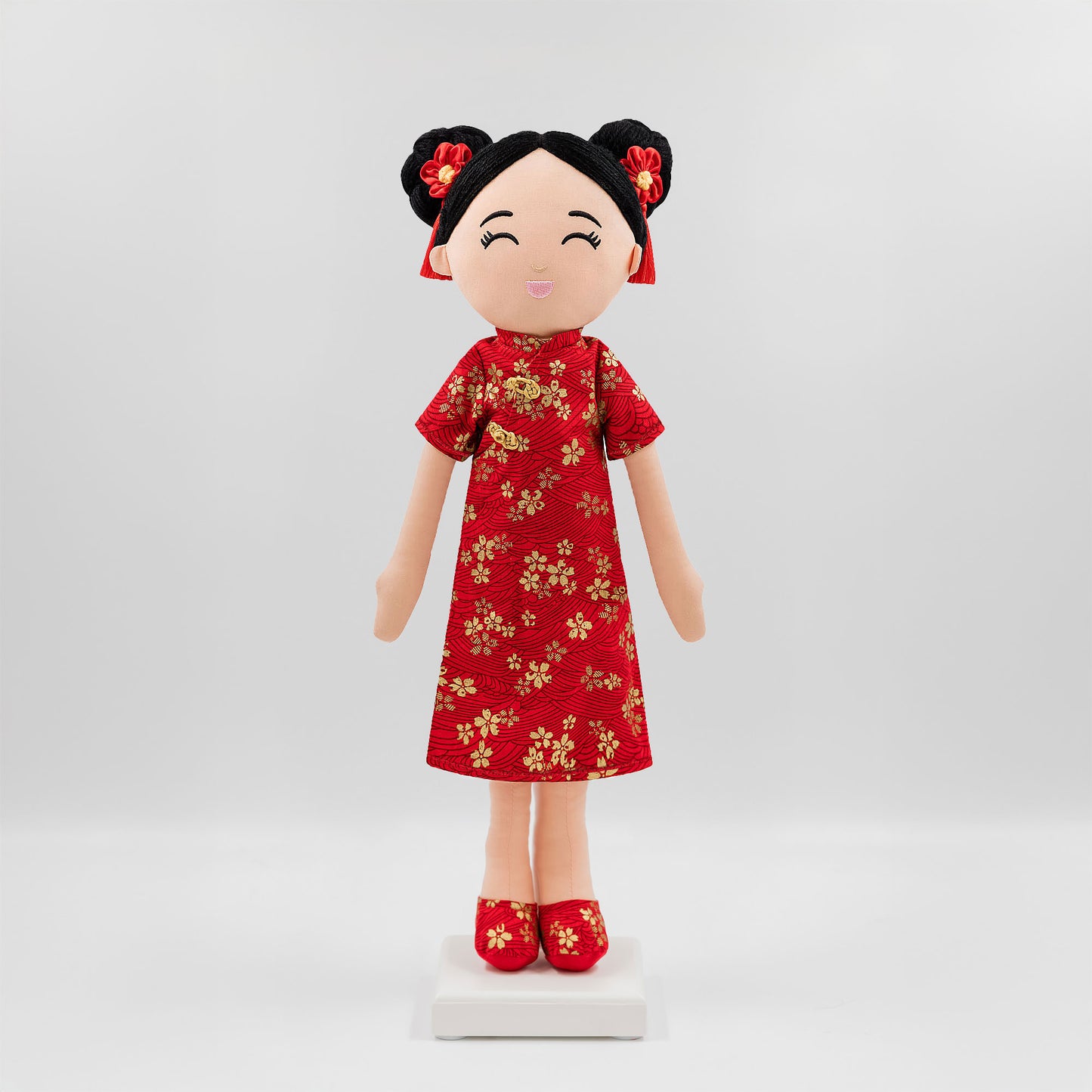 Mei Chinese cultural doll in a front view, wearing a vibrant red qipao adorned with golden floral embroidery with matching red shoes and golden patterns, while her black hair is styled in double buns decorated with red floral tassels.