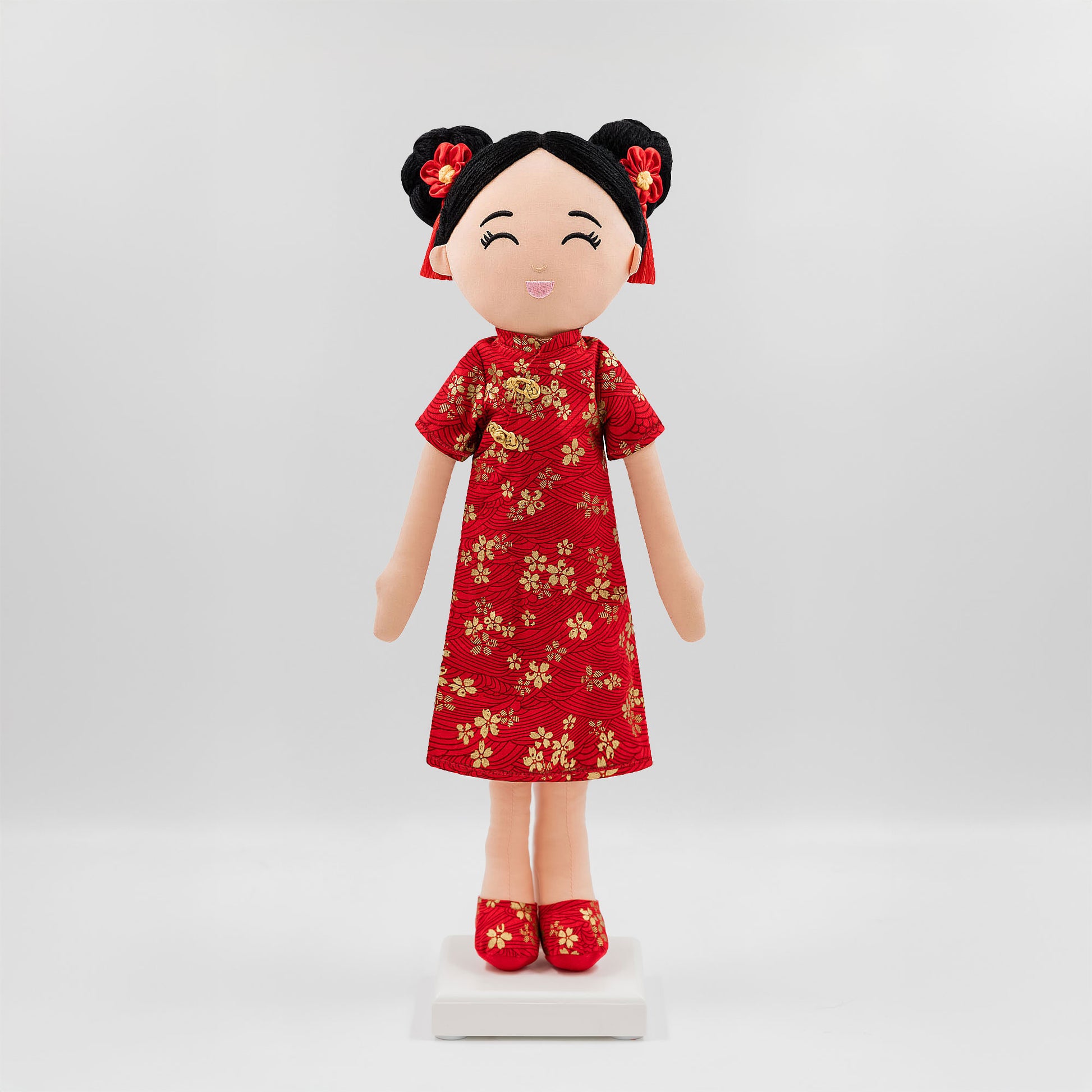 Mei Chinese cultural doll in a front view, wearing a vibrant red qipao adorned with golden floral embroidery with matching red shoes and golden patterns, while her black hair is styled in double buns decorated with red floral tassels.