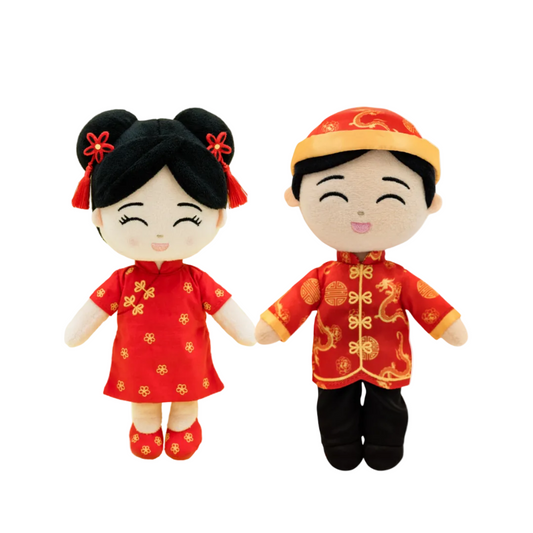 Two plush Chinese New Year dolls standing side by side on a plain white background. The doll on the left is female with black hair in two buns, wearing a red qi pao dress with gold patterns and red shoes.