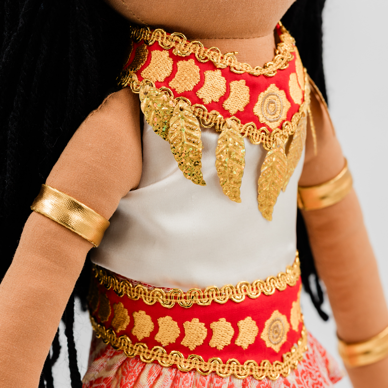 Close-up of Soriya Cambodian cultural doll’s white satin top, featuring an ornate gold and red collar with leaf-shaped sequins and a matching gold-patterned red belt at her waist, complemented by gold armbands on her wrists and arms.