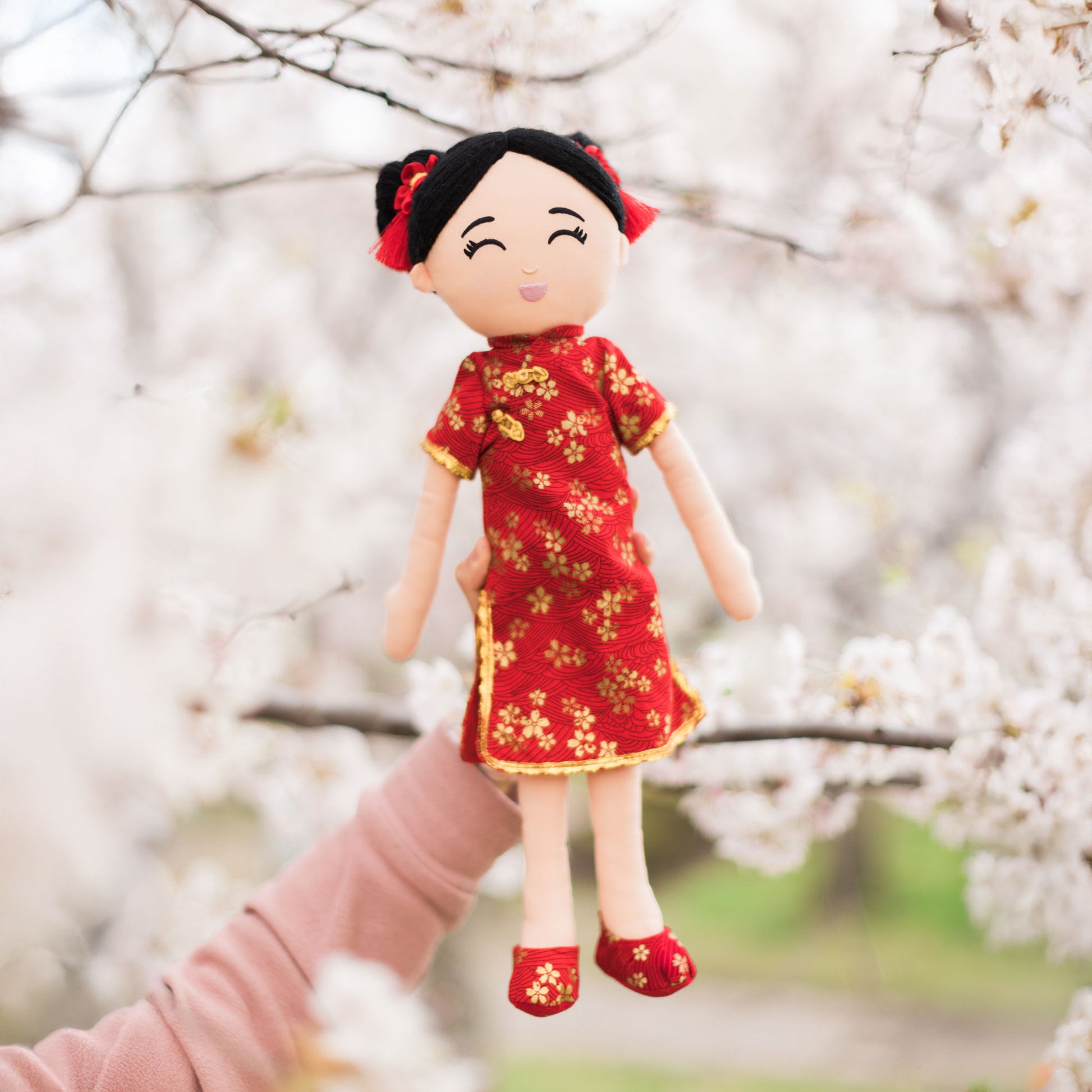 Mei Chinese cultural doll in a front view, beautifully held against a soft background of white cherry blossom trees. 