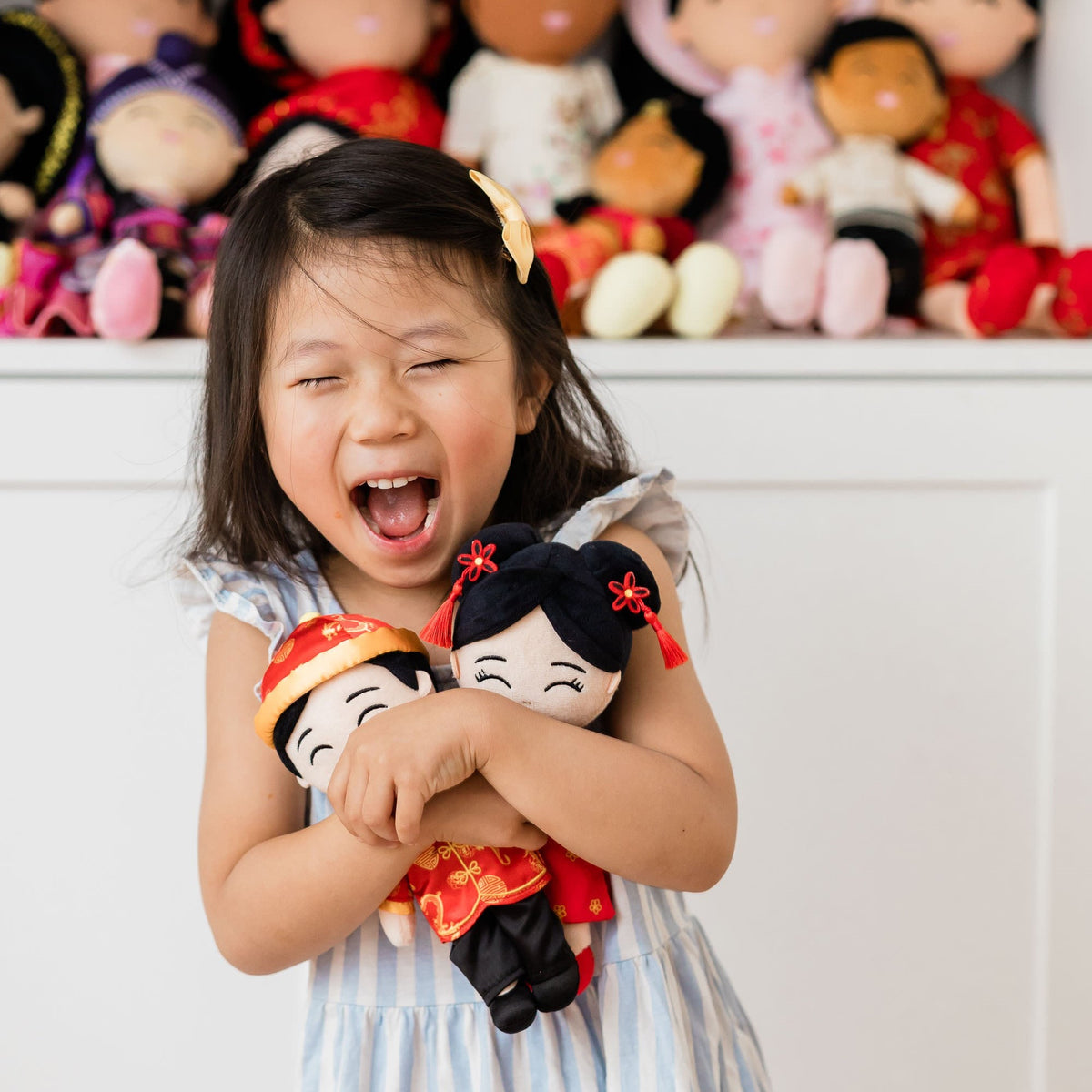 Laughing girl hugging Mei and Jun, Joeydolls Chinese mini dolls, in front of a shelf of cultural dolls