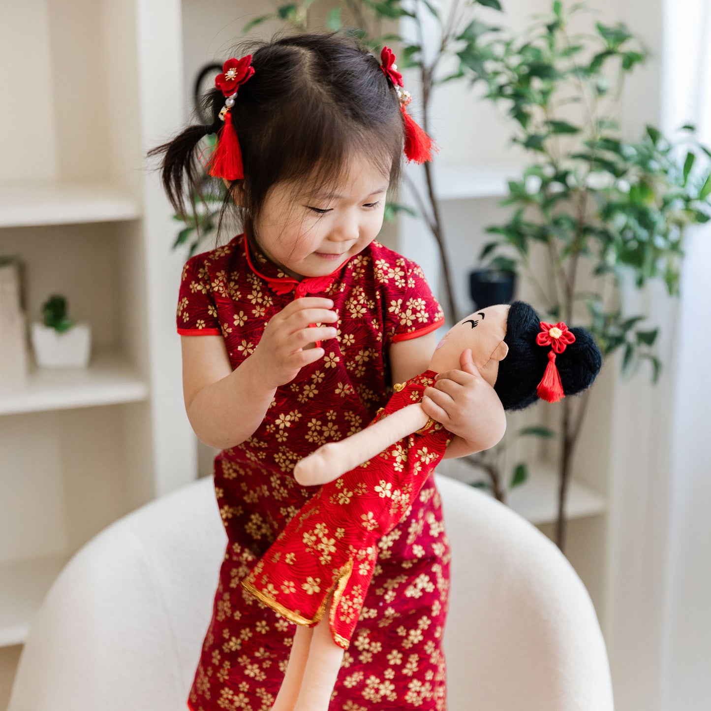 A young girl in a matching red and gold qipao smiling while joyfully holding the Mei Chinese cultural doll. Both the girl and the doll are styled with red floral hair tassels on their black hair.
