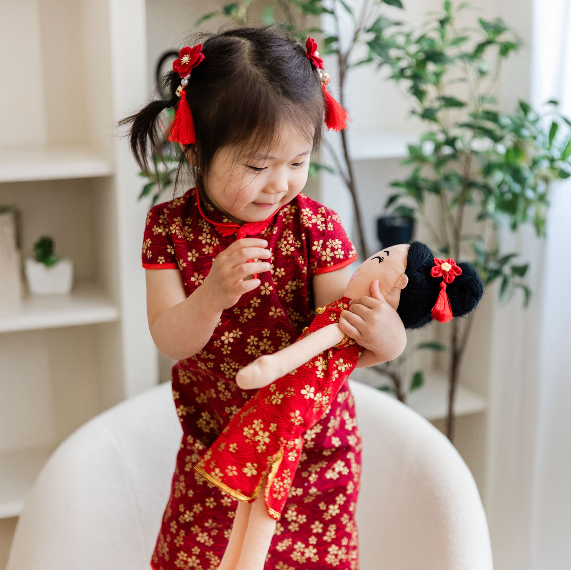 A young girl in a matching red and gold qipao smiling while joyfully holding the Mei Chinese cultural doll. Both the girl and the doll are styled with red floral hair tassels on their black hair.
