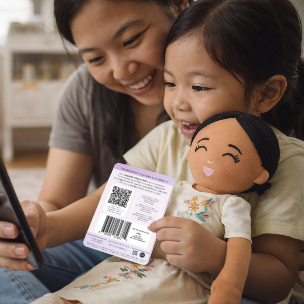 Woman and child holding a doll and a product label, with a blurred indoor background.