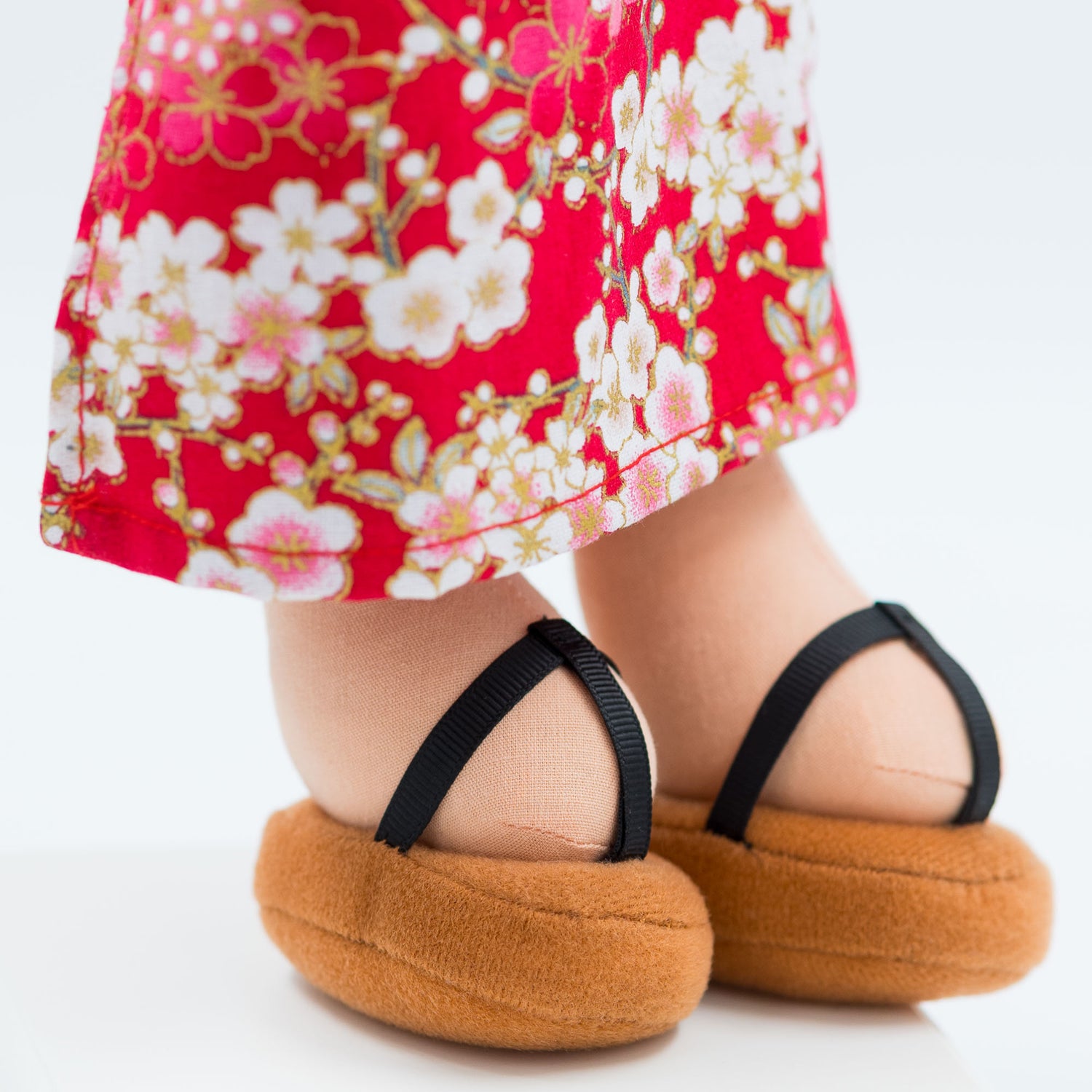 Close-up of Aiko Japanese cultural doll’s lower outfit, showing the hem of her red sakura-patterned kimono over brown plush Zori sandals with black straps.
