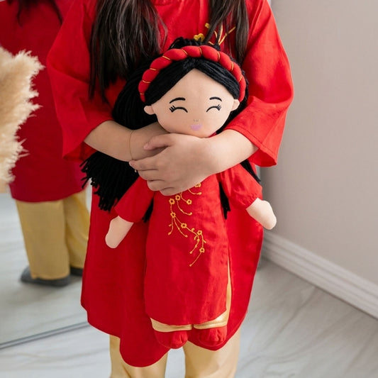 A young girl warmly hugging the Hoa Vietnamese cultural doll to her chest. Both the girl and the doll are dressed in matching vibrant red áo dài and gold trousers, featuring the doll's joyful face with squinting eyes, pink cheeks, and a red braided headband with gold accents on her long black hair.