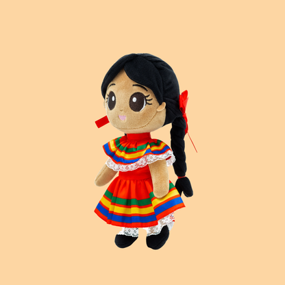 Meet Xochitl 🌸 (pronounced so-cheel), named after the Nahuatl word for “flower.” Left-side view of a plush Mexican girl doll in a vibrant Jalisco folklórico dress with rainbow ribbon details, white lace trim, and braided hair with red ribbons on a peach background.