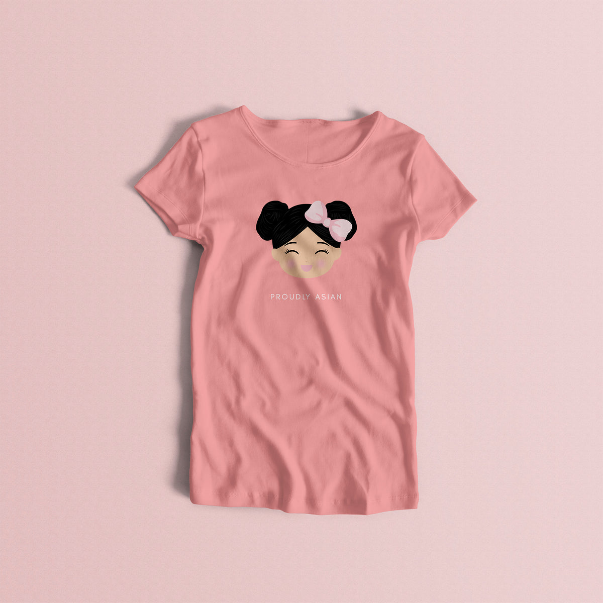 Proudly Asian women’s t-shirt in pink featuring a printed illustration of a smiling girl’s face with black hair in two buns and a bow, with the text “PROUDLY ASIAN” below.