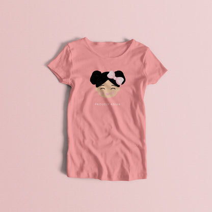 Proudly Asian women’s t-shirt in pink featuring a printed illustration of a smiling girl’s face with black hair in two buns and a bow, with the text “PROUDLY ASIAN” below.