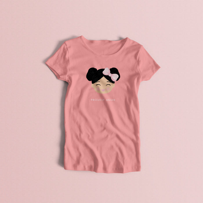 Proudly Asian women’s t-shirt in pink featuring a printed illustration of a smiling girl’s face with black hair in two buns and a bow, with the text “PROUDLY ASIAN” below.