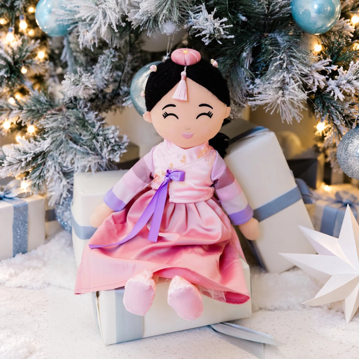 Danbi Korean plush doll in a pink hanbok, seated on a gift beneath a frosted Christmas tree.