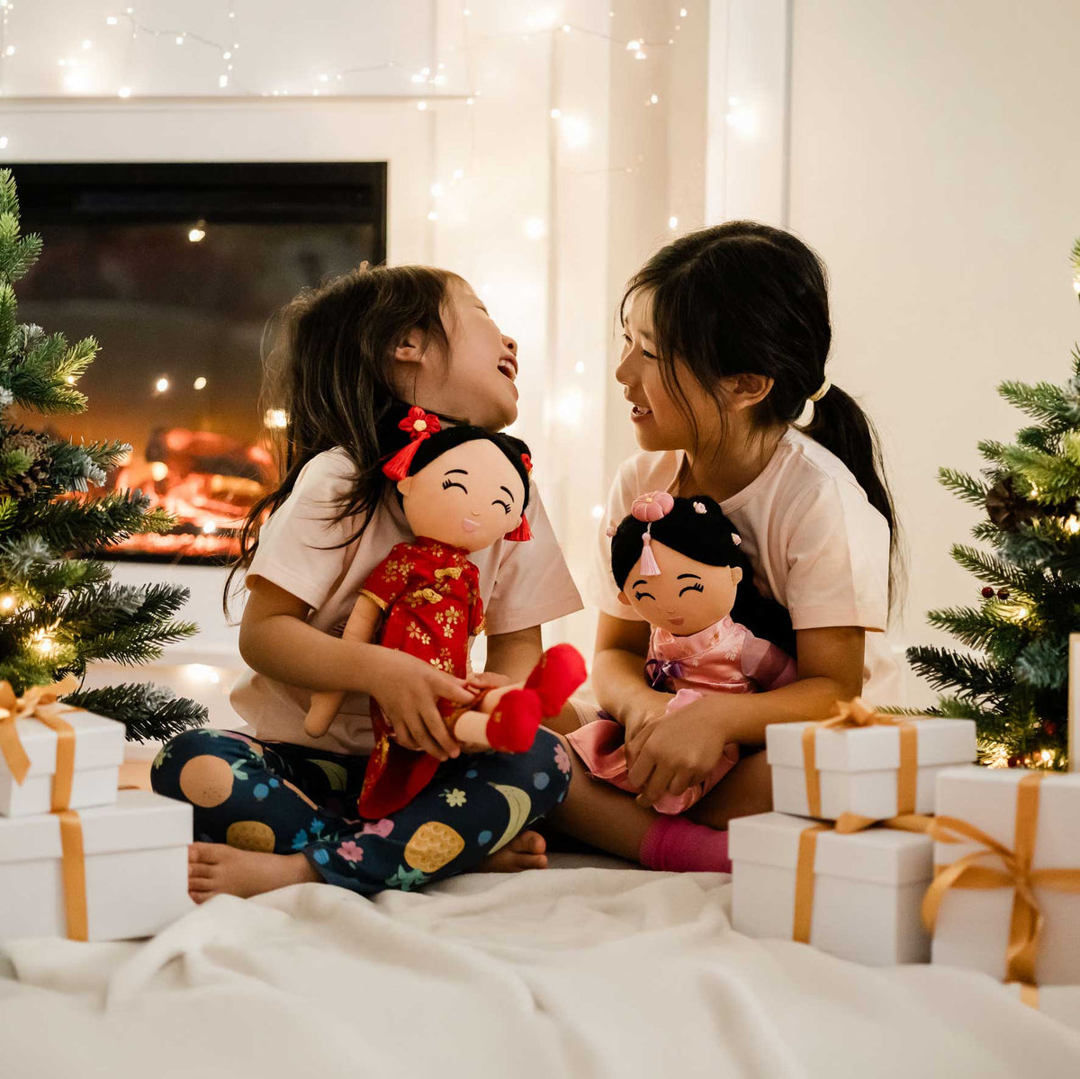 Two laughing children in Cultural Celebration tees hugging Joeydolls Signature dolls by a fireplace and holiday trees