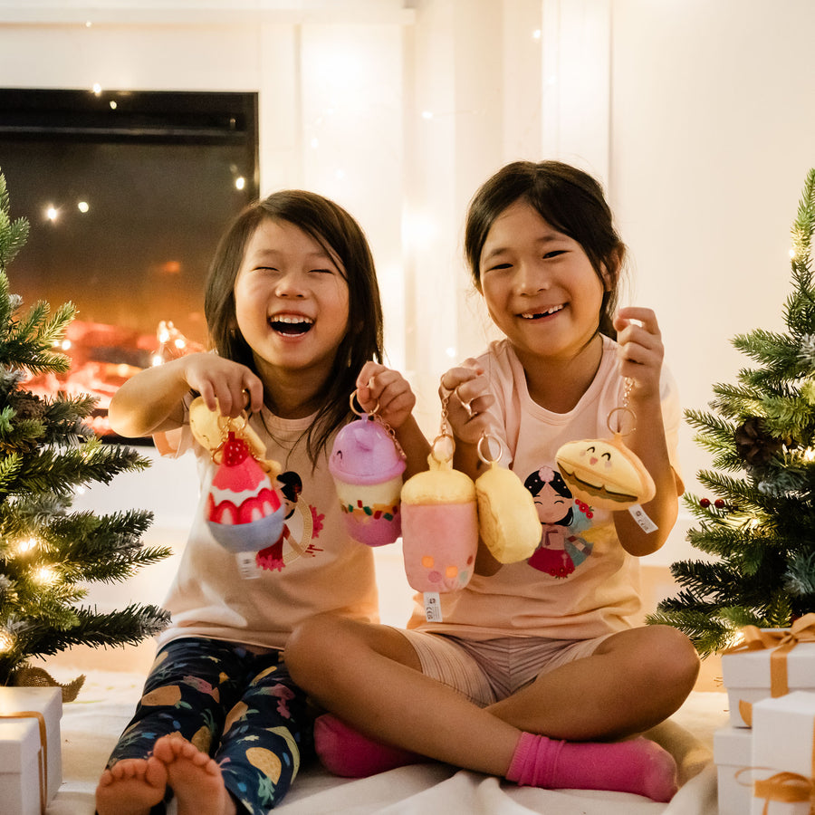 Two smiling children holding up Joeydolls Food Pals plush bag charms beside wrapped gifts