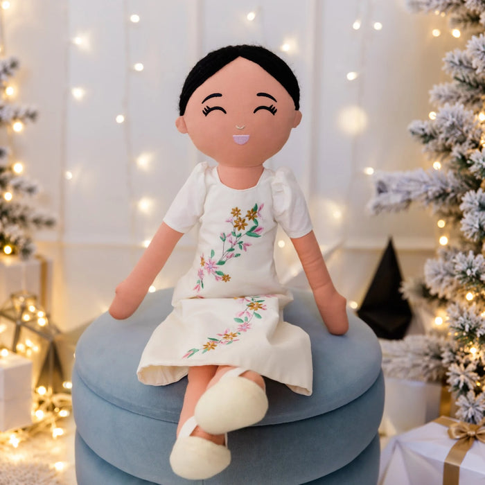 Malaya Filipina plush doll in an ivory embroidered Filipiniana dress, seated on blue cushions with Christmas lights behind her.