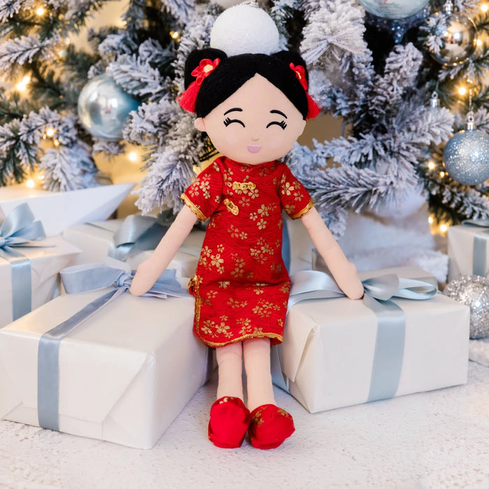 Mei Chinese plush doll in a red and gold floral dress, seated among gifts beneath a frosted Christmas tree.
