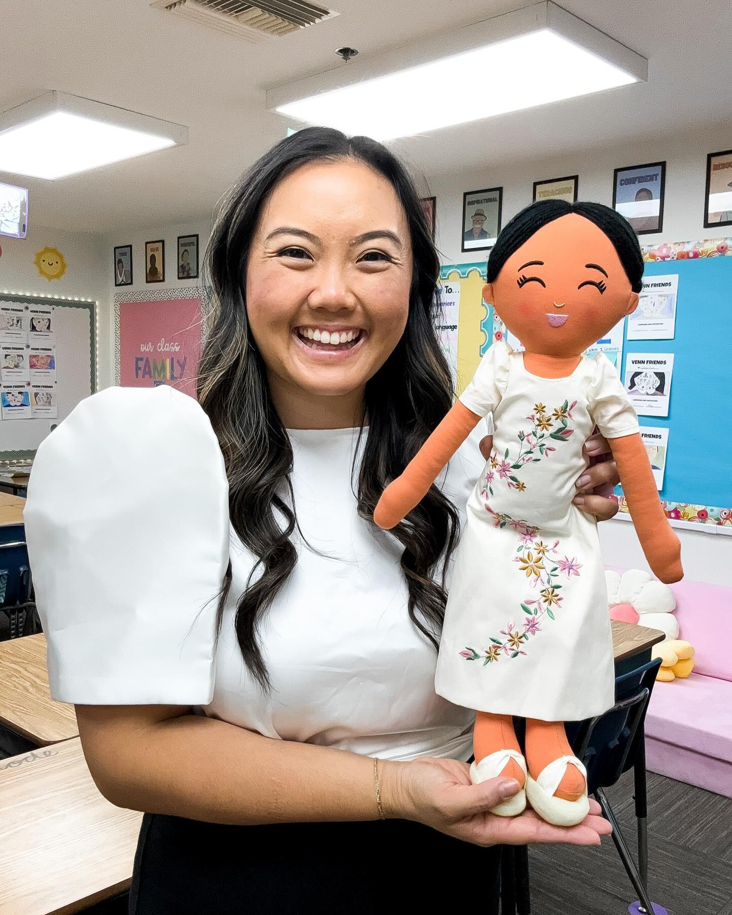 A smiling teacher in a classroom holds a Joeydolls Filipino cultural doll wearing a traditional white embroidered floral dress, highlighting joyful representation and cultural connection through play.